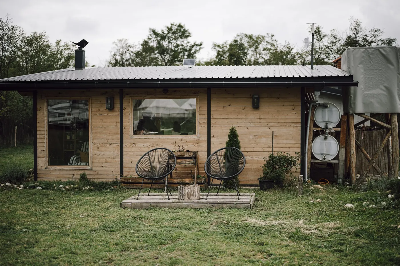 Cabaña de madera con dos sillas en la terraza, rodeada de pasto y árboles